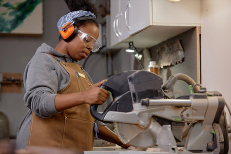 Person focusing intently while using power saw in workshop wearing safety gear including headphones and goggles creating a woodworking project learning carpentry skillsの写真素材