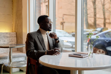 Man sitting by window in cozy cafe, wearing glasses and suit jacket, holding a drink and looking...