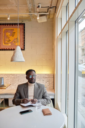 Man wearing sunglasses and beige turtleneck sweater writing notes while sitting at a table by large...