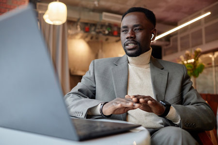 Man in business attire typing on laptop at workspace with modern interior, displaying engagement and professionalism. Includes background elements like plants and lights, creating inviting atmosphereの写真素材