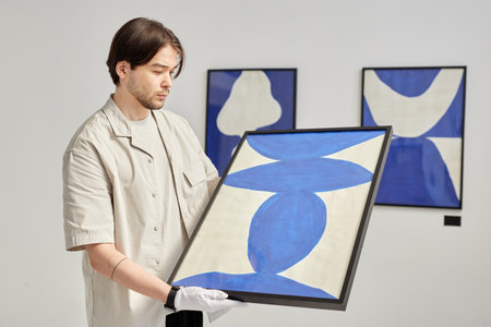 Minimal waist up portrait of young man as curator carefully holding abstract art painting while planning exhibition in modern art gallery copy spaceの写真素材