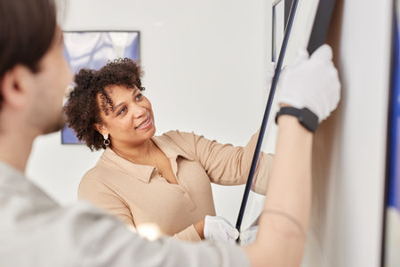 Side view portrait of smiling Black woman hanging painting on wall in modern art gallery or museum with colleagueの写真素材