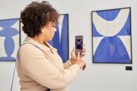 Back view of adult woman holding smartphone and tapping screen while taking pictures or videos of paintings visiting modern art galleryの写真素材