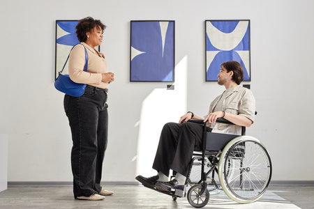 Minimal side view shot of man with disability using wheelchair and talking to friend while visiting accessible art gallery exhibitionの写真素材