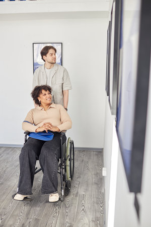 Vertical full length portrait of Black woman with disability using wheelchair in accessible art gallery and looking at paintings with friend or partner assistingの写真素材