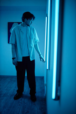 Vertical side view portrait of young man interacting with blue neon light strips during modern art...
