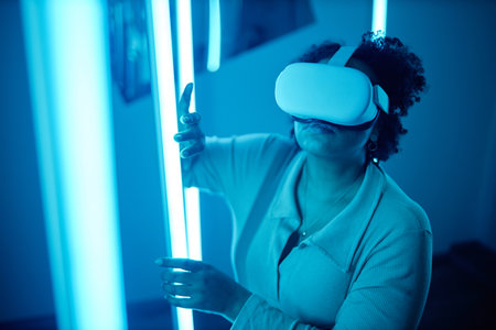 Portrait of African American woman wearing VR headset in modern art show interacting with blue neon...