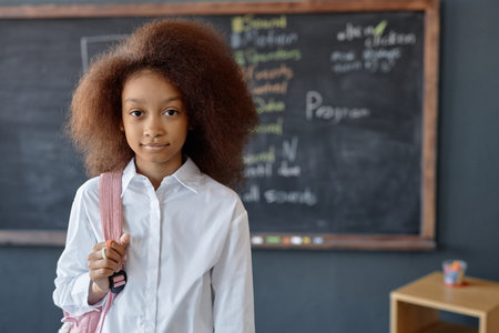 Waist up portrait of cute African American girl with puffy curly hair looking at camera holding...