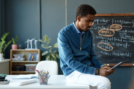 Side view portrait of African American male teacher sitting on desk in school classroom and using...