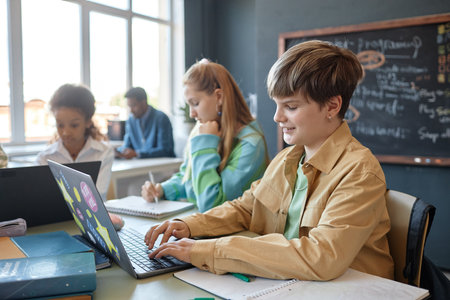 Side view portrait of smiling young boy using laptop while enjoying lesson in modern school...