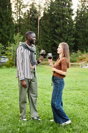 Vertical full length shot of adult couple drinking wine outdoors standing on green lawn and enjoying communicationの写真素材