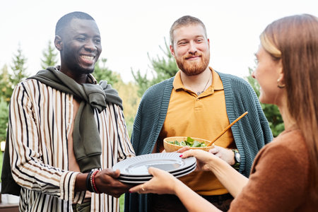 Waist up portrait of group of friends setting dining table during party together and serving dishes focus on African American man smiling happily holding platesの写真素材