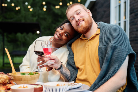 Vibrant portrait of multiethnic adult couple posing for photo sitting at dinner table and holding glasses outdoorsの写真素材