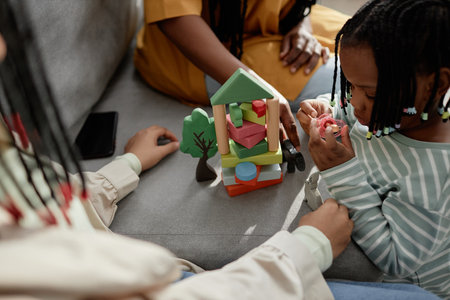 Children sitting and engaging in creative play with a colorful toy. A child in a white shirt is looking at the camera while constructing structures with wooden blocksの写真素材