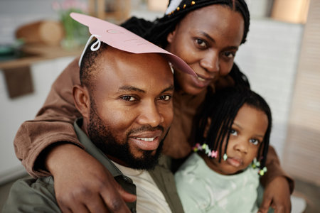 Portrait of smiling family with father wearing paper hat, mother hugging him from behind, and child leaning in closely. All members appearing joyful and relaxed in indoor settingの写真素材