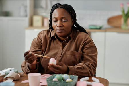 African American woman painting easter eggs in bright kitchen preparing for holiday celebrations...