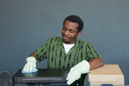 Waist up portrait of smiling African American man cleaning office equipment and wearing gloves on...