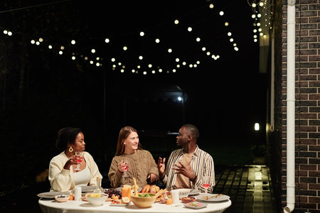 Group of three happy adult people enjoying dinner together outdoors at terrace decorated with...