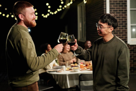 Side view portrait of two adult men clinking glasses and toasting while enjoying party with friends outdoorsの写真素材