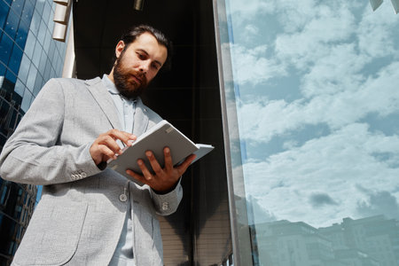 Bearded man analyzing data on tablet while standing outdoors next to glass building on bright day,...