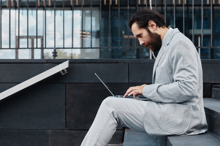 Man with beard wearing gray suit, working on laptop while sitting on outdoor steps. Reflected building glass and modern architecture in background is visibleの写真素材