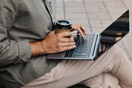 Working person holding coffee cup with laptop placed on lap, outdoors on paved area, focusing on screen without showing faceの写真素材