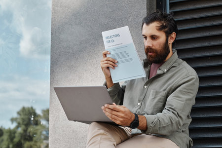 Bearded man sitting outside, holding a paper document, and using a laptop for work, surrounded by...