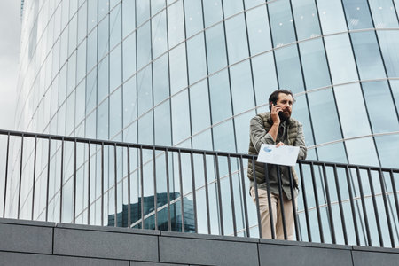 Person standing near modern office building exterior holding smartphone engaged in conversation while holding documents. Surrounded by urban environment and tall glass structuresの写真素材