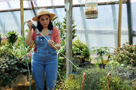 Woman wearing hat watering plants in greenhouse with focused expression holding watering hose among...