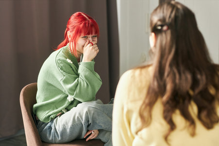 Woman with red hair, wearing green sweater and jeans, smiling while sitting on chair. Blurred person with long hair in foreground, creating focus on the red-haired womanの写真素材