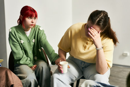 Woman with red hair sitting beside a friend who is covering face with hand, likely offering support...