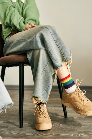 Person seated on chair wearing rainbow socks and beige sneakers with crossed legs, casual denim...