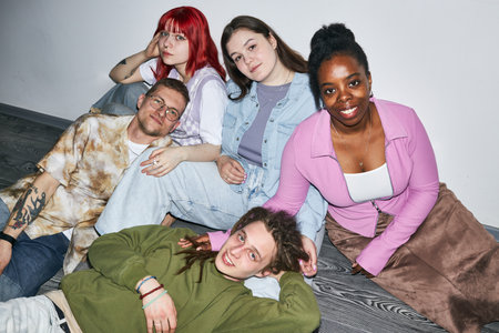 Group of friends sitting on floor smiling at camera, showcasing diverse backgrounds. They are...