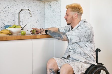 Caucasian man with tattoos peeling apple in modern kitchen setting, using wheelchair for mobility and independenceの写真素材