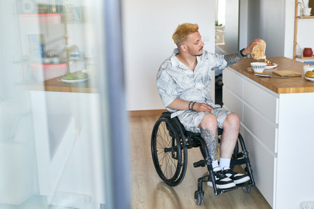 Man in wheelchair placing coffee cup on kitchen counter with baked goods in background and modern kitchen design. Space for design on white cabinets and countertopの写真素材