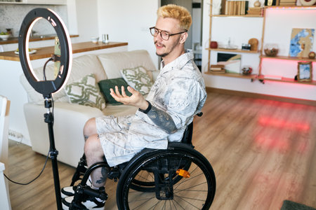 Man in wheelchair recording educational video in modern living room setting using ring light and camera wearing glasses and speaking with confidenceの写真素材