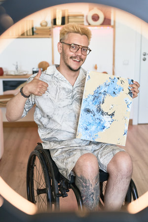 Smiling man in wheelchair holding abstract painting in home setting enthusiastically. Man with artistic tattoo showing his creativity while sitting in decorated living roomの写真素材