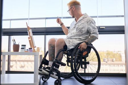 Man with tattoos, sitting in wheelchair, painting on easel in bright room with large windows and city view, creating art piece in casual attire with focus on creativity and concentrationの写真素材