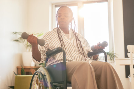 African American woman in wheelchair lifting dumbbells with focused expression in brightly lit room during day. Plant and shelf visible in background creating a homely atmosphereの写真素材
