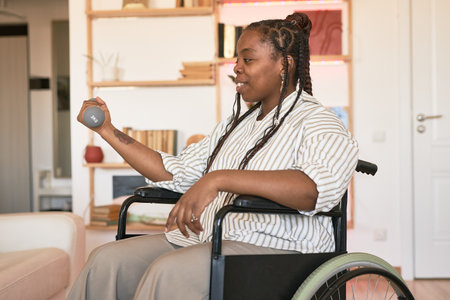 Black person sitting in wheelchair and exercising with dumbbell in hand while surrounded by home...