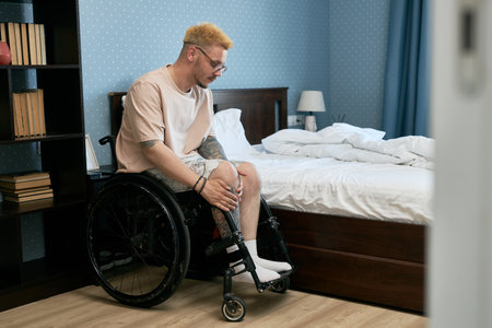 Man sitting in wheelchair adjusting prosthetic leg beside unmade bed in cozy bedroom. Bookshelf filled with various books visible in backgroundの写真素材