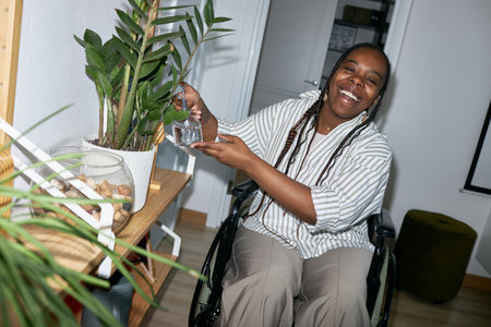 African American woman in wheelchair smiling while watering potted plant with a watering can. She is...