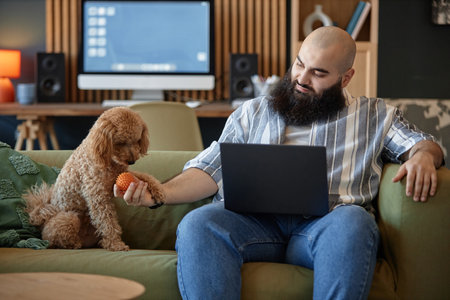 Bearded man engaging with brown dog while sitting on couch with laptop in living room filled with...