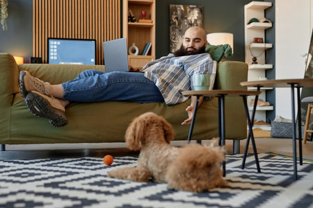 Man reclining on couch with laptop on lap, working remotely in cozy living room featuring modern furniture, bookshelf, and adorable dogの写真素材