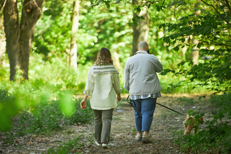 Couple walking along forest trail, holding hands and enjoying nature hike with their dog leashed beside them, surrounded by lush green foliage and tranquilityの写真素材