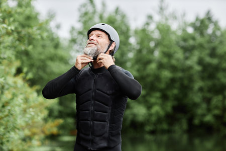 Waist up portrait of bearded senior man putting on wetsuit and helmet ready to enjoy water sports by lake in forest copy spaceの写真素材