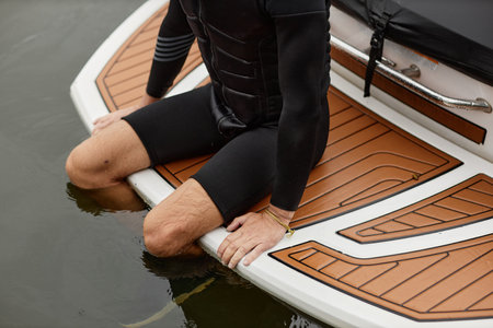 Cropped shot of unrecognizable sportive man wearing wetsuit sitting on back of boat with legs in...