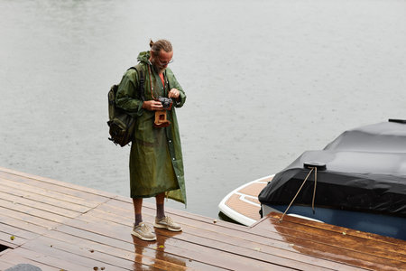 Candid wide angle shot of male tourist wearing raincoat and holding vintage photo camera while standing by water on wooden pier copy spaceの写真素材