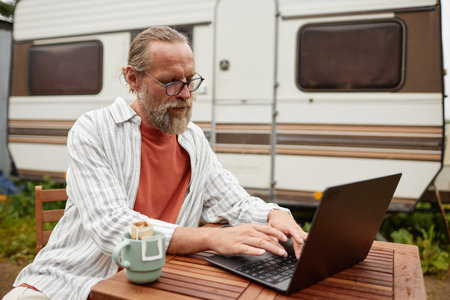 Side view portrait of bearded senior man working with laptop outdoors sitting by van, copy spaceの写真素材