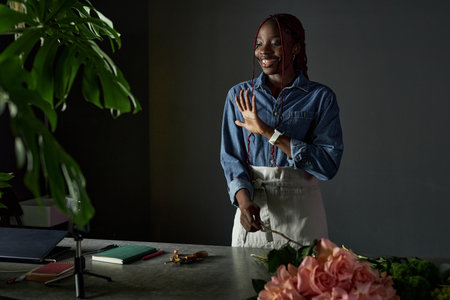 Minimal portrait of young African American woman as florist creating bouquet and filming process with smartphone for social media waving at camera copy spaceの写真素材